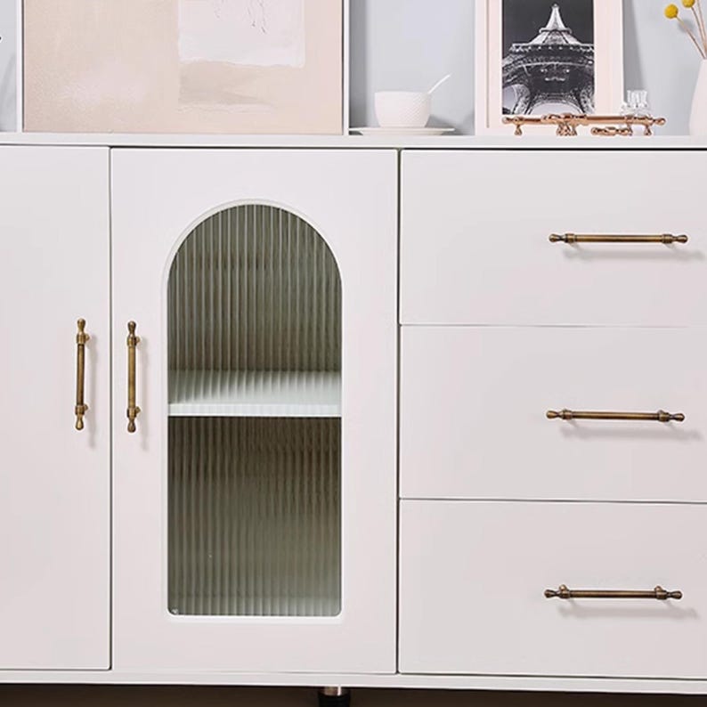May include: A white cabinet with a glass door and three drawers. The cabinet has brass-coloured handles and a shelf inside the glass door. The glass door has a ribbed texture. The cabinet is part of a furniture set.