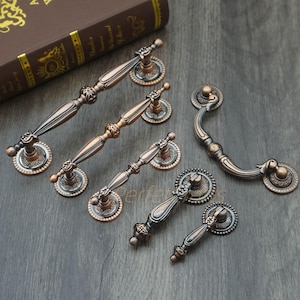 Könnte beinhalten: Eine Auswahl an antiken bronzenen Schrankgriffen und -griffen. Die Hardware weist verzierte Designs mit einer Mischung aus dunklen und hellen Bronzefarben auf. Die Griffe variieren in Größe und Stil, mit dekorativen Rückplatten und detaillierten Griffen.