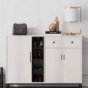 May include: A white shoe cabinet with black detailing, including a shoe rack, drawers, and doors. A black quilted handbag, gold ornaments, and a lamp with a white shade are on top. The cabinet rests on a patterned rug.