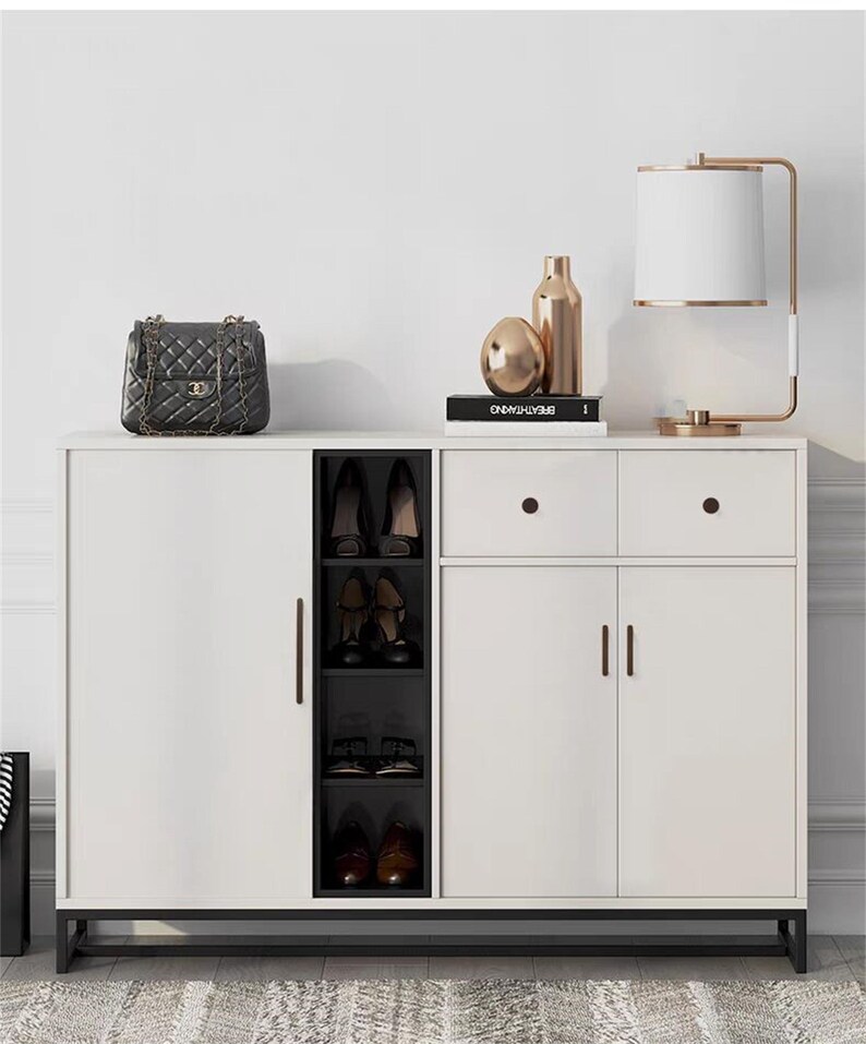 May include: A white shoe cabinet with black detailing, including a shoe rack, drawers, and doors. A black quilted handbag, gold ornaments, and a lamp with a white shade are on top. The cabinet rests on a patterned rug.