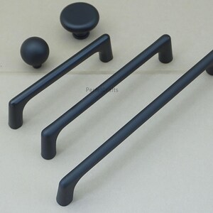 May include: A collection of matte black cabinet hardware, including two knobs and three bar pulls. The knobs are round, and the pulls have a curved design. The hardware is displayed on a neutral background.