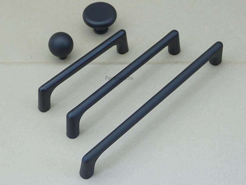 May include: A collection of matte black cabinet hardware, including two knobs and three bar pulls. The knobs are round, and the pulls have a curved design. The hardware is displayed on a neutral background.