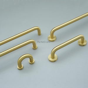 May include: A selection of brass-toned cabinet pulls and handles in different sizes. The hardware has a curved design with circular mounting plates. The finish is a matte gold, and the items are displayed against a neutral grey backdrop.