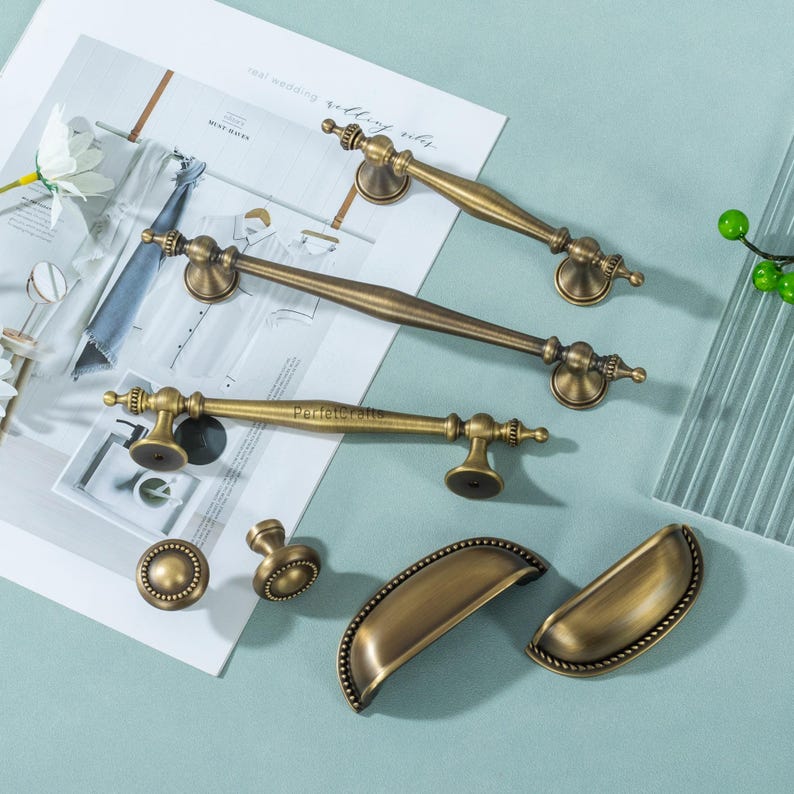 Może przedstawiać: Asortyment antycznych mosiężnych okuć do szafek, w tym uchwyty, gałki i uchwyty. Uchwyty r&oacute;żnią się długością, z dekoracyjnymi detalami na końcach. Okucia ułożone są na jasnoniebieskiej powierzchni, z magazynem i kwiatowymi akcentami w tle.