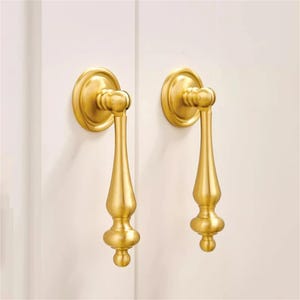 Może przedstawiać: Dwa złote uchwyty do szafek o klasycznym designie. Każdy uchwyt posiada okrągłą płytkę tylną i dekoracyjny, wydłużony uchwyt z zaokrąglonym dnem. Uchwyty są zamontowane na białej powierzchni.