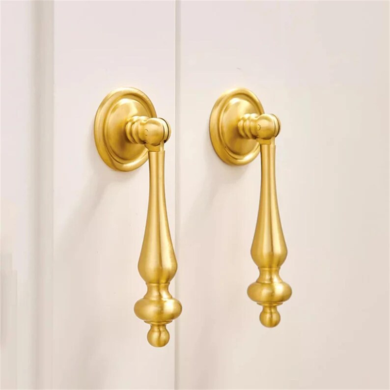 Może przedstawiać: Dwa złote uchwyty do szafek o klasycznym designie. Każdy uchwyt posiada okrągłą płytkę tylną i dekoracyjny, wydłużony uchwyt z zaokrąglonym dnem. Uchwyty są zamontowane na białej powierzchni.