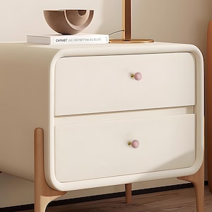 May include: Cream-coloured bedside table with two drawers and pink knobs. The table has rounded edges and sits on light-coloured wooden legs. A decorative bowl and a book are on top.