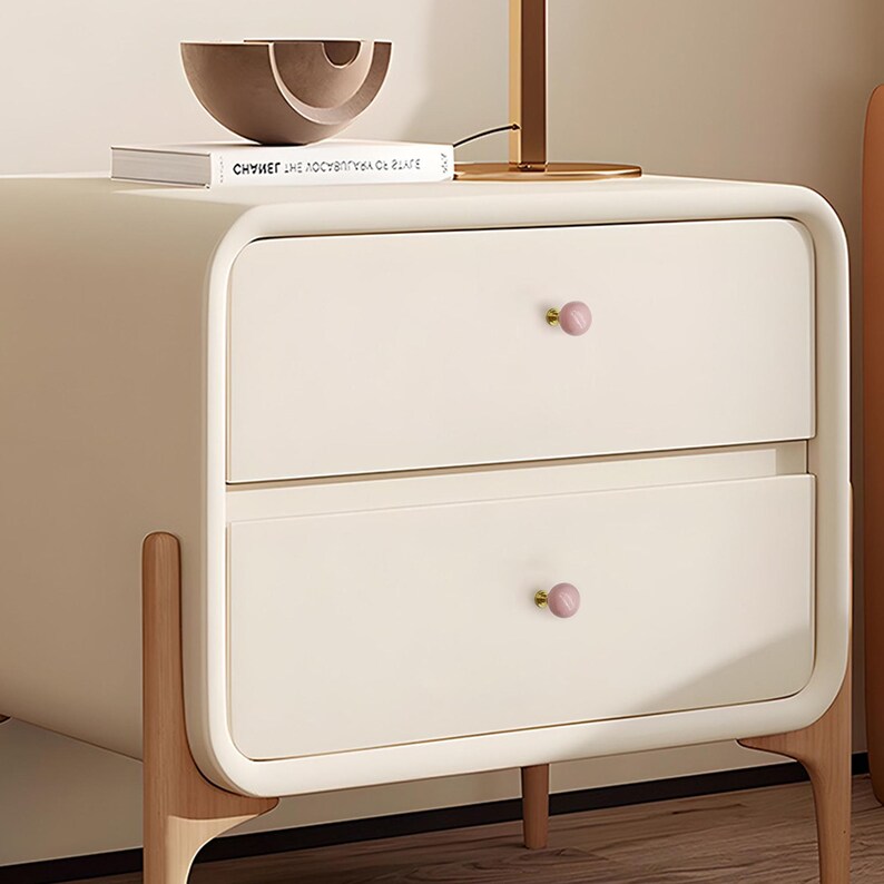 May include: Cream-coloured bedside table with two drawers and pink knobs. The table has rounded edges and sits on light-coloured wooden legs. A decorative bowl and a book are on top.