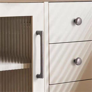 May include: A close-up of a white cabinet with a glass door and drawers. The cabinet has a dark gray handle and round knobs. The glass door has vertical lines, and the drawers are plain. The cabinet is made of wood.
