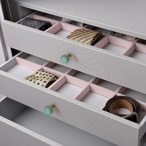 May include: A grey wooden chest of drawers with open drawers, displaying neatly arranged ties and a belt. The drawers have pink dividers and light blue and gold knobs. The ties are various colours and patterns.
