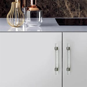 Peut inclure: Design d'int&eacute;rieur moderne avec deux poign&eacute;es de placard en verre transparent avec des accents m&eacute;talliques argent&eacute;s. Les poign&eacute;es sont mont&eacute;es sur une porte d'armoire blanche. Des vases d&eacute;coratifs dor&eacute;s et cuivr&eacute;s sont sur le comptoir.