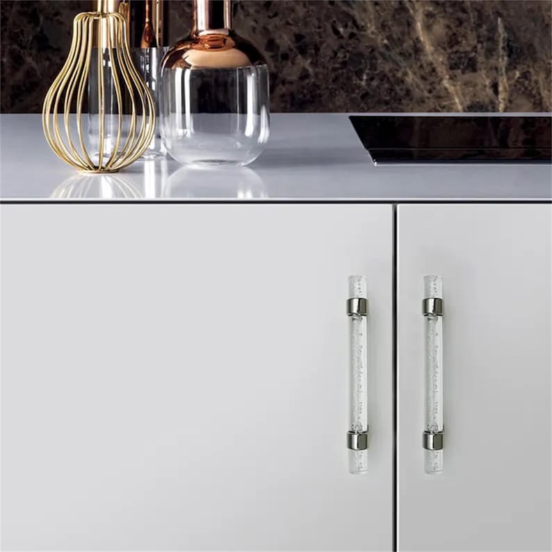 Peut inclure: Design d'int&eacute;rieur moderne avec deux poign&eacute;es de placard en verre transparent avec des accents m&eacute;talliques argent&eacute;s. Les poign&eacute;es sont mont&eacute;es sur une porte d'armoire blanche. Des vases d&eacute;coratifs dor&eacute;s et cuivr&eacute;s sont sur le comptoir.