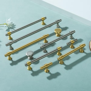 May include: A selection of cabinet handles and knobs in silver and gold tones. The handles are bar-shaped with gold detailing, and the knobs are round. The hardware is displayed on a light blue surface, showcasing different sizes and styles for home decor, in cm.