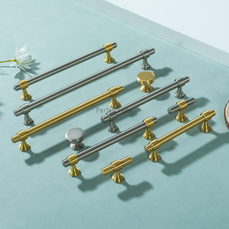May include: A selection of cabinet handles and knobs in silver and gold tones. The handles are bar-shaped with gold detailing, and the knobs are round. The hardware is displayed on a light blue surface, showcasing different sizes and styles for home decor, in cm.