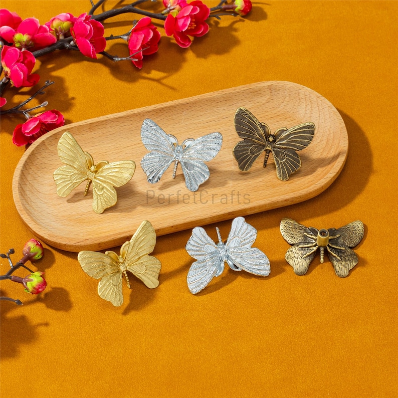 May include: Six butterfly-shaped cabinet knobs in gold, silver, and bronze tones are arranged on a wooden tray. The knobs are detailed with intricate wing patterns and are set against a warm, orange background, with red flowers in the top left corner.