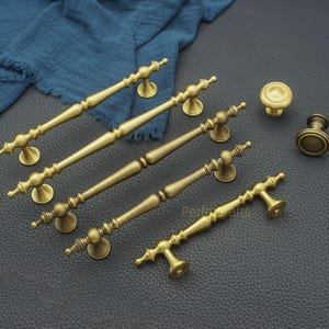 May include: A collection of antique brass cabinet hardware, including several pull handles and two knobs. The handles vary in size and feature ornate detailing. The hardware is displayed on a dark surface with a blue cloth in the background.