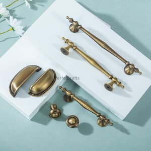 Może przedstawiać: Asortyment antycznych mosiężnych okuć do szafek, w tym uchwyty, uchwyty i gałki. Okucia charakteryzują się bogatymi detalami i ciepłym, postarzanym wykończeniem. Elementy ułożone są na białej powierzchni z jasnoniebieskim tłem.