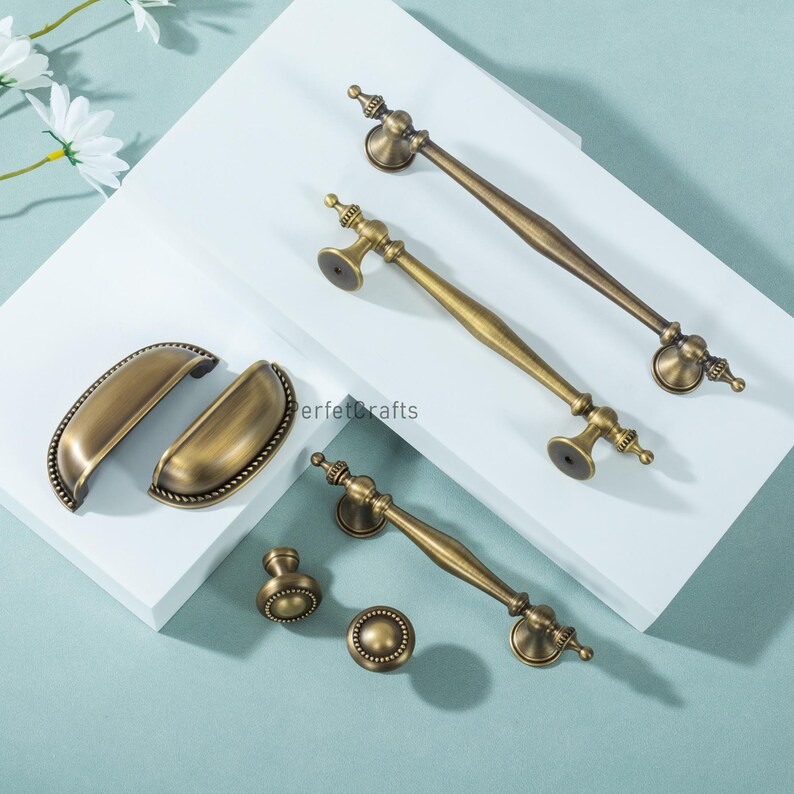 Może przedstawiać: Asortyment antycznych mosiężnych okuć do szafek, w tym uchwyty, uchwyty i gałki. Okucia charakteryzują się bogatymi detalami i ciepłym, postarzanym wykończeniem. Elementy ułożone są na białej powierzchni z jasnoniebieskim tłem.