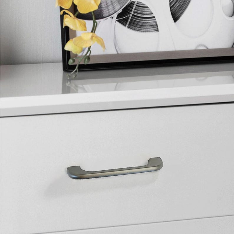 Op de afbeelding: Close-up van een witte kastlade met een handgreep van geborsteld metaal. De handgreep heeft de vorm van een gebogen staaf. Een ingelijste zwart-wit print en gele bloemen zijn zichtbaar op de achtergrond.