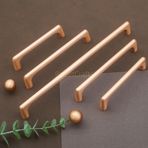 Op de afbeelding: Een verzameling koperkleurige kastgrepen en knoppen. De grepen zijn staafvormig met gebogen uiteinden en de knoppen zijn bolvormig. De hardware is gerangschikt op een bruin oppervlak met een takje groen.