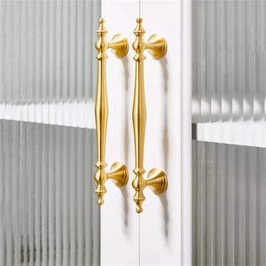 Może przedstawiać: Dwa złote uchwyty do drzwi szafy. Każdy uchwyt ma długi, smukły korpus z dekoracyjnymi detalami i jest zamontowany na białych drzwiach szafy. Uchwyty są symetryczne i wydają się być wykonane z metalu.