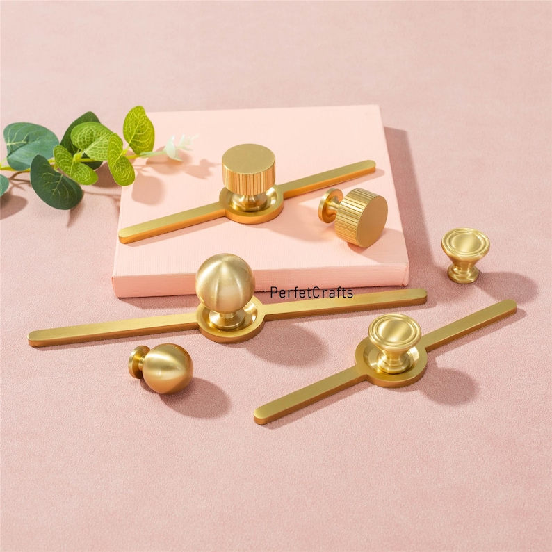 Puede incluir: Una colecci&oacute;n de herrajes dorados para armarios, que incluyen pomos y tiradores, dispuestos sobre una superficie rosa. Los herrajes presentan varios dise&ntilde;os, incluyendo elementos cil&iacute;ndricos, esf&eacute;ricos y en forma de disco. El texto "PerfektCrafts" es visible.