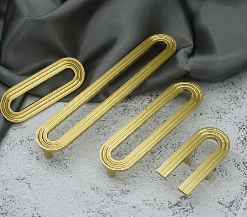 May include: A selection of gold-toned cabinet handles in different sizes. The handles feature a curved, elongated shape with vertical grooves. They are displayed on a grey surface with a draped fabric backdrop.