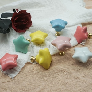 Può includere: Una collezione di pomelli per mobili a forma di stella in vari colori, tra cui giallo, rosa, blu e bianco. Ogni pomello ha una base color oro. I pomelli sono disposti su un tessuto bianco con una rosa rossa sullo sfondo. Il testo "PerfetcCrafts" è visibile.