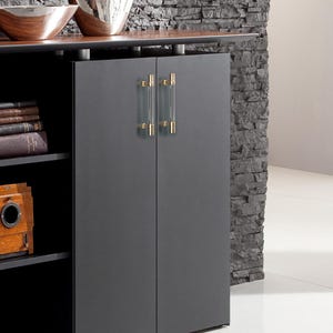 May include: A dark gray cabinet with two doors and gold-toned handles. The cabinet is part of a larger piece of furniture with shelves and decorative bowls. The background features a stone wall.