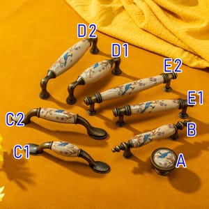 May include: A collection of antique-style cabinet hardware, including handles and a knob. The hardware features a cream-colored ceramic center with a blue bird and floral design, set against a bronze-toned metal base. The handles vary in size and shape.