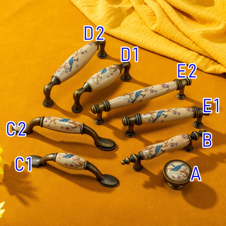 May include: A collection of antique-style cabinet hardware, including handles and a knob. The hardware features a cream-colored ceramic center with a blue bird and floral design, set against a bronze-toned metal base. The handles vary in size and shape.