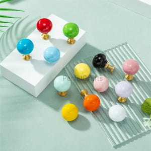 May include: A selection of colourful, spherical cabinet knobs. The knobs are in various colours, including red, green, blue, yellow, orange, pink, black, and white. Each knob has a gold-coloured base.