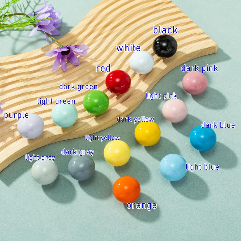May include: A selection of brightly coloured, spherical beads in shades such as black, white, red, and orange. The beads are displayed on a wooden board with colour labels. Purple flowers are also visible in the image.