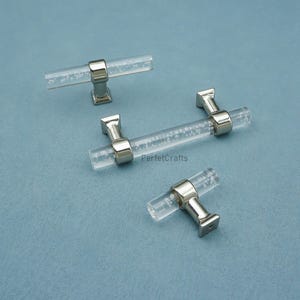 Peut inclure: Quatre poign&eacute;es de meubles en acrylique transparent avec des accents m&eacute;talliques argent&eacute;s. Les poign&eacute;es ont une forme cylindrique avec de petites bulles &agrave; l'int&eacute;rieur de l'acrylique. Elles sont dispos&eacute;es sur une surface bleue. Le texte "PerfetcCrafts" est visible.