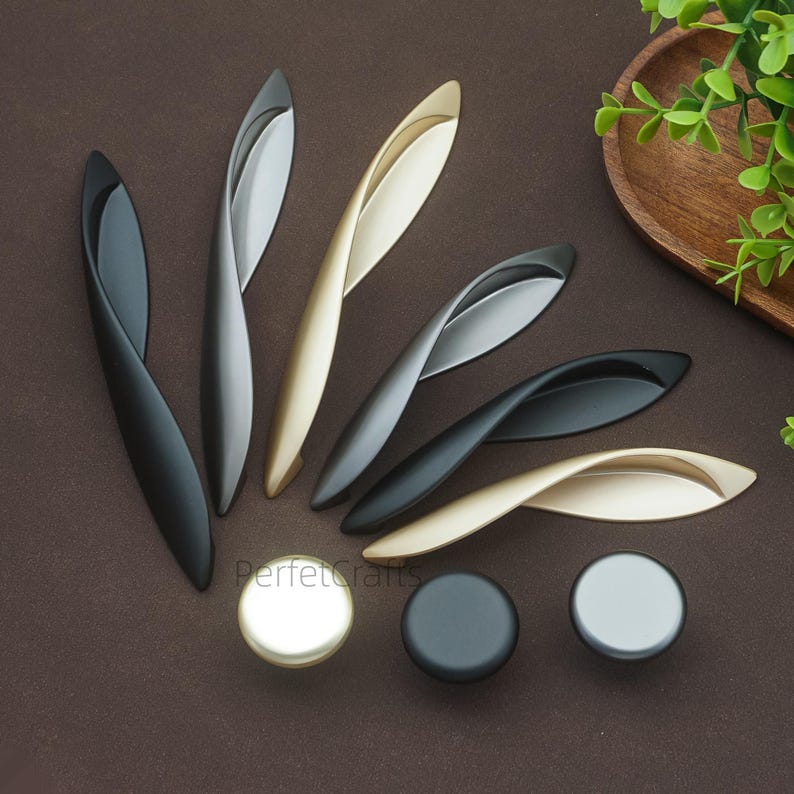 以下が含まれることがあります： さまざまな金属仕上げのモダンなキャビネットハードウェアのコレクション。 黒、シルバー、ゴールドトーンの曲線ハンドルと、お揃いの丸いノブが含まれています。 ハードウェアは茶色の表面に配置されています。