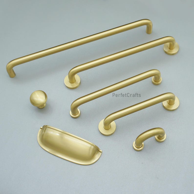May include: A selection of gold-coloured cabinet hardware, including bar pulls, a knob, and a cup pull. The hardware has a matte finish and is arranged on a light grey surface. The design is modern and minimalist, suitable for various furniture styles.