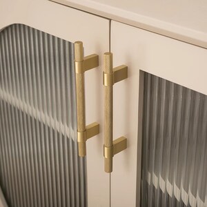 K&ouml;nnte beinhalten: Nahaufnahme einer Schrankt&uuml;r mit zwei goldfarbenen, strukturierten Griffen. Die Schrankt&uuml;r ist cremefarben mit einer vertikalen, geriffelten Glasscheibe. Die Griffe sind zylindrisch mit einem strukturierten Muster und rechteckigen Montageplatten.