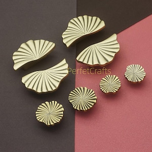 Può includere: Una collezione di pomelli e maniglie per mobili color oro. I pomelli sono rotondi con un design a raggiera, mentre le maniglie sono a forma di ventaglio con una texture a coste. Lo sfondo è un mix di marrone e rosa.