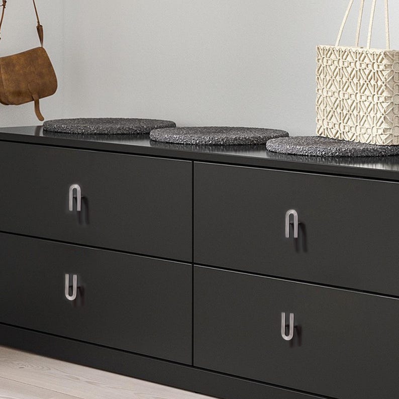 May include: A black chest of drawers with four drawers and silver U-shaped handles. The top has three dark grey, textured decorative mats. A woven white tote bag and a brown leather bag hang in the background.