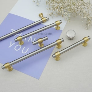 Peut inclure: Ensemble de quincaillerie d'armoires, comprenant des poign&eacute;es et un bouton argent et or. Les poign&eacute;es ont un design cylindrique avec des accents dor&eacute;s. Le mot "YOU" est visible sur un fond violet. La marque "PerfecCrafts" est &eacute;galement visible.