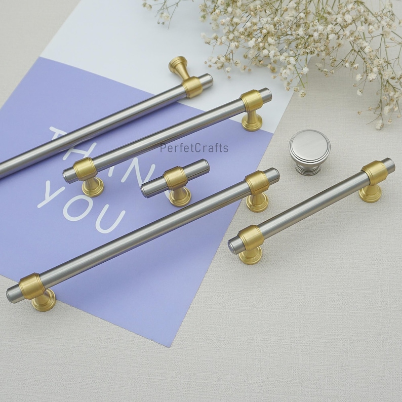 Peut inclure: Ensemble de quincaillerie d'armoires, comprenant des poign&eacute;es et un bouton argent et or. Les poign&eacute;es ont un design cylindrique avec des accents dor&eacute;s. Le mot "YOU" est visible sur un fond violet. La marque "PerfecCrafts" est &eacute;galement visible.