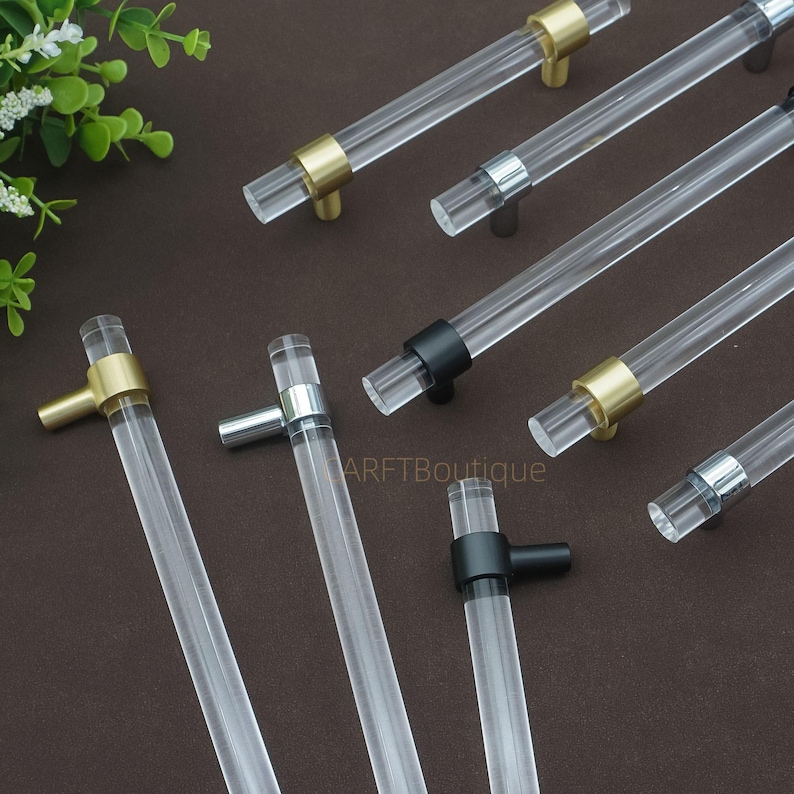 May include: Clear acrylic cabinet pulls with gold, silver, and black metal finishes. The cylindrical pulls are displayed on a brown surface. These handles are suitable for drawers and cabinets. The image shows a variety of designs.