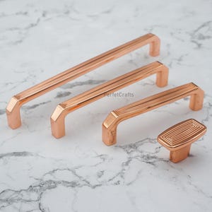 May include: Four copper-colored cabinet hardware pieces. The set includes three handles of varying lengths and a single knob. Each piece features a linear grooved design and a rectangular base. The hardware is displayed on a marble surface.