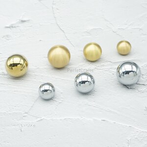 Op de afbeelding: Een assortiment van bolvormige kastknoppen in verschillende maten en afwerkingen. De knoppen omvatten gepolijst messing, geborsteld messing en chroom, die licht reflecteren. De knoppen zijn gerangschikt op een getextureerd wit oppervlak. De tekst "PerfetcCrafts" is zichtbaar.