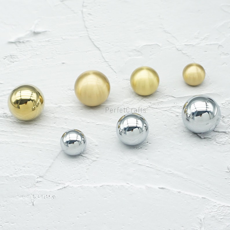 Op de afbeelding: Een assortiment van bolvormige kastknoppen in verschillende maten en afwerkingen. De knoppen omvatten gepolijst messing, geborsteld messing en chroom, die licht reflecteren. De knoppen zijn gerangschikt op een getextureerd wit oppervlak. De tekst "PerfetcCrafts" is zichtbaar.
