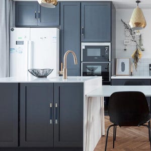 Puede incluir: Una cocina moderna con gabinetes azul oscuro, una encimera blanca y un grifo dorado. Se ven un refrigerador blanco y un microondas y horno empotrados. Una silla negra est&aacute; sentada en una mesa blanca. Una l&aacute;mpara colgante dorada cuelga del techo.