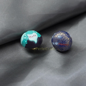 May include: Two decorative planet-themed knobs. One is teal and purple with "Pluto" in white. The other is blue with gold accents and "Mercury" in red. The knobs are likely for furniture or decor.