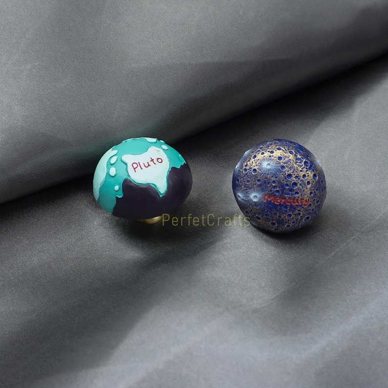 May include: Two decorative planet-themed knobs. One is teal and purple with "Pluto" in white. The other is blue with gold accents and "Mercury" in red. The knobs are likely for furniture or decor.