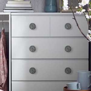 May include: White dresser with six decorative knobs. Each knob is round with a gray and black pattern and red text. The dresser is in a room with a vase, books, and a decorative birdcage.