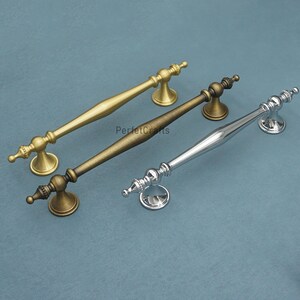 Może przedstawiać: Trzy dekoracyjne uchwyty do szafek w kolorze złotym, brązowym i srebrnym. Każdy uchwyt ma ozdobny design z zakrzywionym prętem i dekoracyjnymi końc&oacute;wkami. Uchwyty są prezentowane na niebiesko-zielonej powierzchni. Widoczny jest tekst "PerfectCrafts".